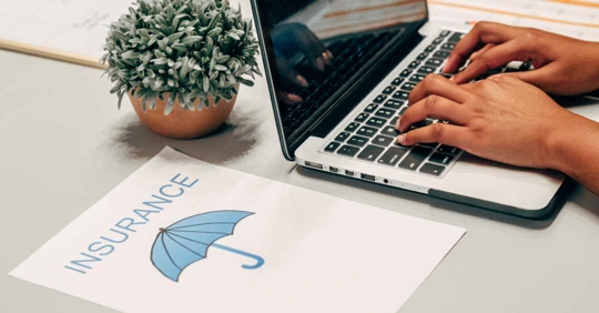 Someone typing on a computer next to a piece of paper that reads "insurance" with a blue umbrella on it