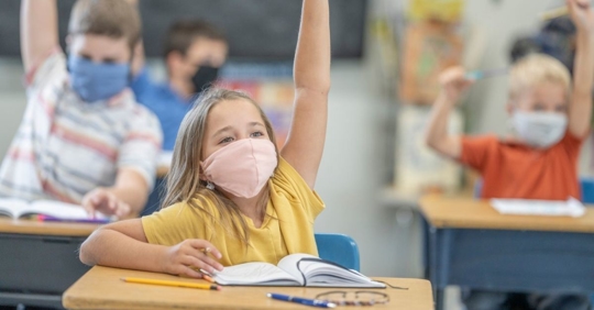 A young girl wearing a mask raises her hand in class to answer the teacher's question