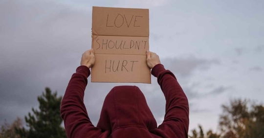 Someone wearing a hoodie holding a sign that reads "Love shouldn't hurt."