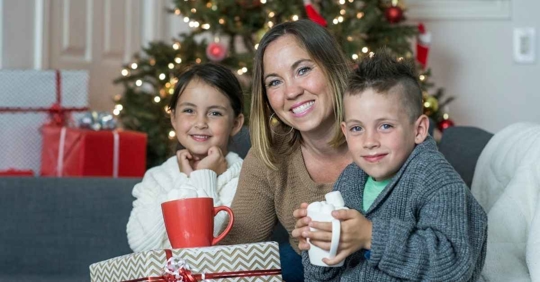 A mother, her son, and her daughter sitting on a couch in front of a holiday tree. The mother is holding a present, while the son is holding a mug of cocoa