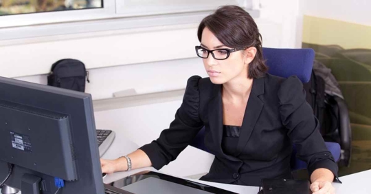 A woman sitting at a desk looking at a computer. She is wearing glasses and a black sport coat