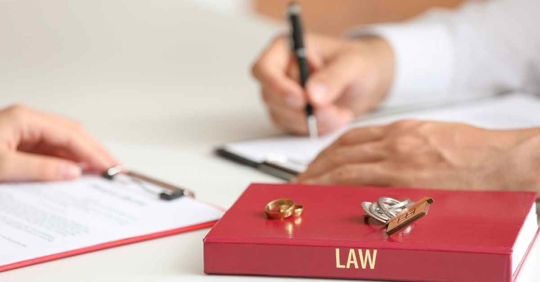 Two people sitting at a desk writing on clipboards. A book reading "law" rests near them, and two wedding rings are on top of the book