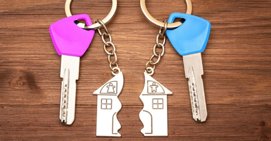 Blue and pink colored keys with split house matching keychains on a wooden table. Splitting assets in a divorce.