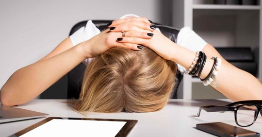 A woman with her head against a desk. She is holding her hands on top of her head. She appears frustrated.