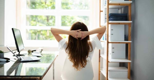 A woman looking out her home office window. Her hands are behind her head, and she appears relaxed or contemplative
