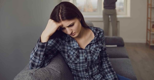 A woman sitting on a couch frustrated. Someone is in the background looking out a window