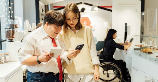 A person with Downs syndrome looking at their co-worker's phone. In the background, a woman in a wheelchair is working at a computer