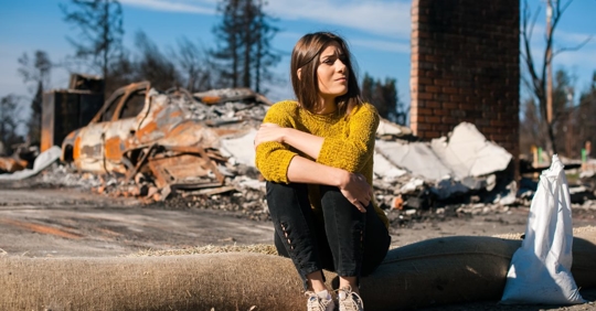 Woman sitting in front of a destroyed home and car